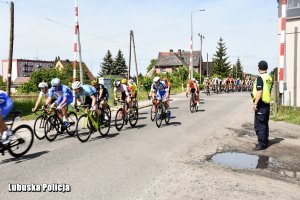 Kolarze przejeżdżają przez przejazd kolejowy. W pobliżu stoi policjant uczestniczący w zabezpieczeniu.