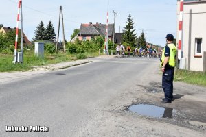 Peleton zbliża się do przejazdu kolejowego. Na poboczu stoi policjant.